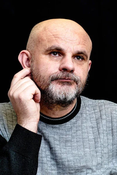 middle-aged bald bearded man touches his ear on the dark background