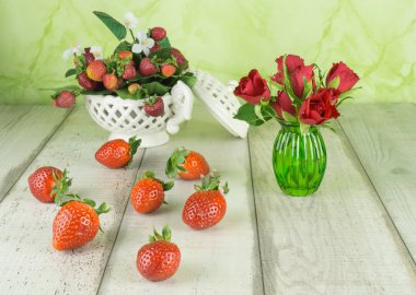 Taze çilek beyaz bir kase ve tahta masada yanında küçük bir yeşil cam vazoda kırmızı gül çiçekler