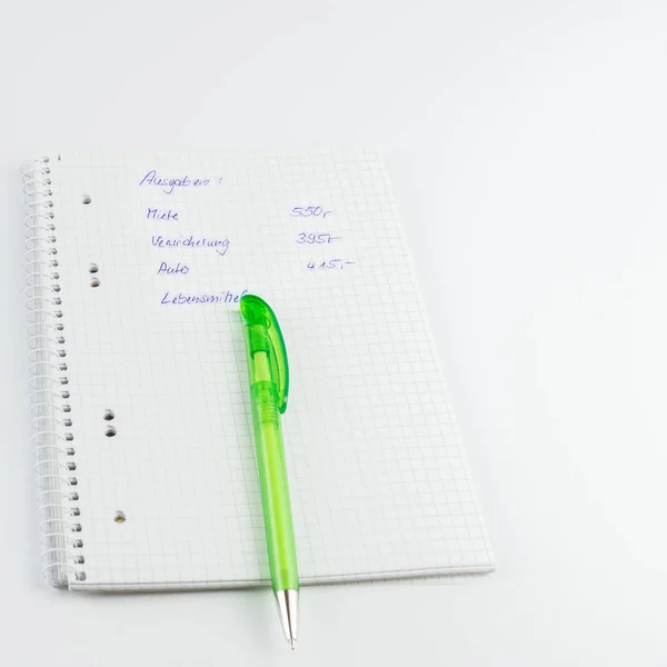 A green ballpoint pen and a writing pad with a calculation of expenses, German words for expenses, rent, insurance, car, food