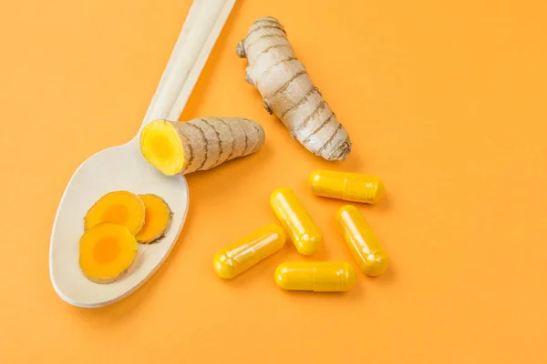 Turmeric root with curcuma capsules and wooden spoon on orange background