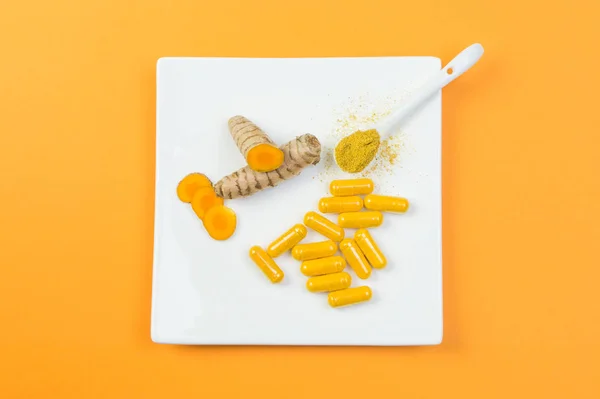 Turmeric root and curcuma capsules and a spoonful curcuma powder on a white plate, orange color background