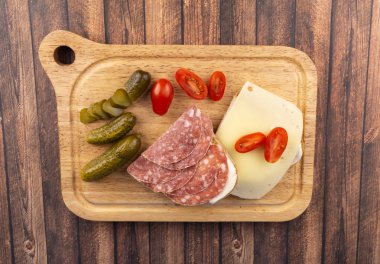 Bir dilim salamlı ekmek ve peynirli bir dilim ekmek, hala küçük domates ve salatalık turşusu, ahşap bir tahtada servis ediliyor.