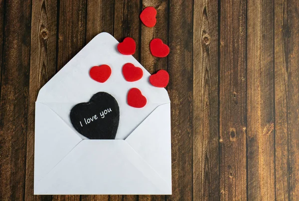 A white envelope with a black heart and the words I love you and red hearts, wooden table as a background