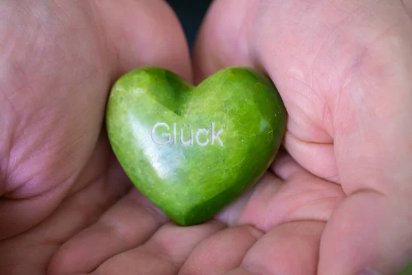 Holding a green heart of stone in the hand, in white letters the German word for luck