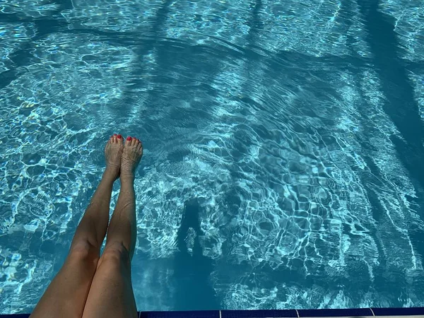 Pieds dans la piscine. Détendez-vous, vacances, été. Ongles rouges, peau bronzée, eau turquoise. La lumière du soleil danse dans l'eau. Vacances d'été, détente, plaisir, bronzage. Flottant à la surface — Photo