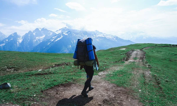 A male traveler in the mountains with a huge backpack on his shoulders. The traveler is walking along the road, and against the background of the tops of snow-capped mountains.