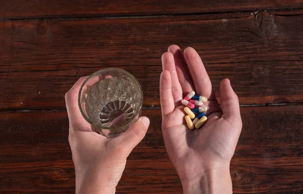 Hold pills and a glass of water in your hand. Wash down the vitamins with clean, clear water. A glass of water in one hand and pills in the other. Wooden background and vitamins.