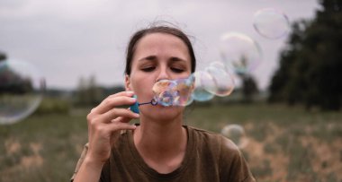 Kız sabun köpüğü üflüyor. Genç bir kadın doğada oturur ve sabun toplarını emer. Öndeki kişi..