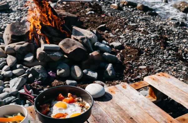 Fry eggs on a gas burner against the background of a fire and sea waves. Delicious and healthy Breakfast in nature on the beach. Fried eggs with tomatoes in a pan.