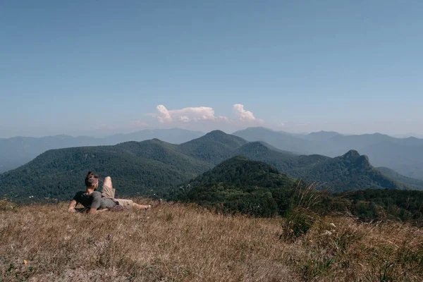 Bir erkek gezgin bir tepenin tepesinde oturur ve etrafındaki dağlara bakar. Güzel dağ ve tepe manzarası sıcak, güneşli hava ve genç turist.