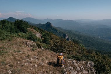 Dişi bir gezgin bir tepenin tepesinde oturur ve etrafındaki dağlara bakar. Güzel dağ ve tepe manzarası sıcak, güneşli hava ve genç turist.