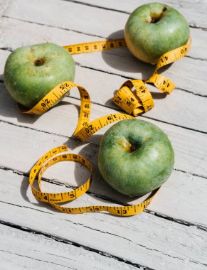 Beyaz ahşap bir zeminde üç yeşil elma uzanır ve onların yanında sarı esnek metre uzunluğunda bir kurdele vardır. Sağlıklı bir yaşam tarzı için yiyecek, lezzetli ve sağlıklı yiyecekler, vitaminler ve mineraller..