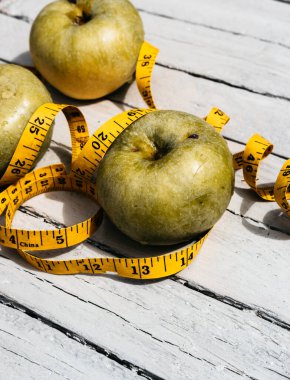 Beyaz ahşap bir zeminde üç yeşil elma uzanır ve onların yanında sarı esnek metre uzunluğunda bir kurdele vardır. Sağlıklı bir yaşam tarzı için yiyecek, lezzetli ve sağlıklı yiyecekler, vitaminler ve mineraller..