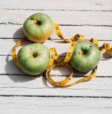 Beyaz ahşap bir zeminde üç yeşil elma uzanır ve onların yanında sarı esnek metre uzunluğunda bir kurdele vardır. Sağlıklı bir yaşam tarzı için yiyecek, lezzetli ve sağlıklı yiyecekler, vitaminler ve mineraller..