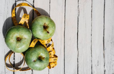 Beyaz ahşap bir zeminde üç yeşil elma uzanır ve onların yanında sarı esnek metre uzunluğunda bir kurdele vardır. Sağlıklı bir yaşam tarzı için yiyecek, lezzetli ve sağlıklı yiyecekler, vitaminler ve mineraller..