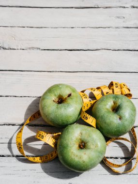 Beyaz ahşap bir zeminde üç yeşil elma uzanır ve onların yanında sarı esnek metre uzunluğunda bir kurdele vardır. Sağlıklı bir yaşam tarzı için yiyecek, lezzetli ve sağlıklı yiyecekler, vitaminler ve mineraller..