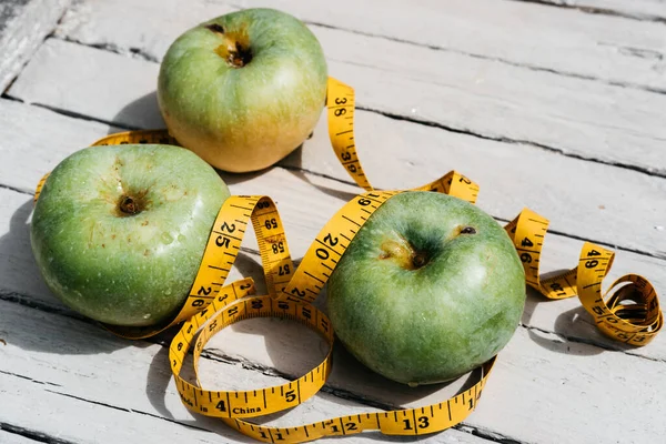 Beyaz ahşap bir zeminde üç yeşil elma uzanır ve onların yanında sarı esnek metre uzunluğunda bir kurdele vardır. Sağlıklı bir yaşam tarzı için yiyecek, lezzetli ve sağlıklı yiyecekler, vitaminler ve mineraller..