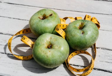 Beyaz ahşap bir zeminde üç yeşil elma uzanır ve onların yanında sarı esnek metre uzunluğunda bir kurdele vardır. Sağlıklı bir yaşam tarzı için yiyecek, lezzetli ve sağlıklı yiyecekler, vitaminler ve mineraller..