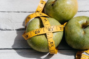 Beyaz ahşap bir zeminde üç yeşil elma uzanır ve onların yanında sarı esnek metre uzunluğunda bir kurdele vardır. Sağlıklı bir yaşam tarzı için yiyecek, lezzetli ve sağlıklı yiyecekler, vitaminler ve mineraller..