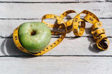 Esnek sarı bir banda sarılı yeşil bir Apple beyaz bir ahşap masanın üzerinde yatıyor. Fitness ve sağlıklı yaşam tarzı. Lezzetli ve sağlıklı yiyecekler, vitaminler ve mineraller.