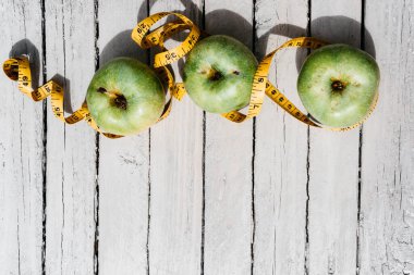 Beyaz ahşap bir zeminde üç yeşil elma uzanır ve onların yanında sarı esnek metre uzunluğunda bir kurdele vardır. Sağlıklı bir yaşam tarzı için yiyecek, lezzetli ve sağlıklı yiyecekler, vitaminler ve mineraller..