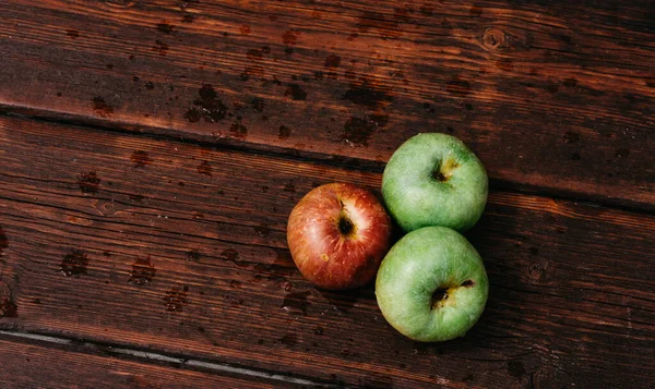Three apples - two green and one red-lie on a wooden table. Fresh apples on a wood textured brown background, mahogany material. Fresh apples, autumn harvest.