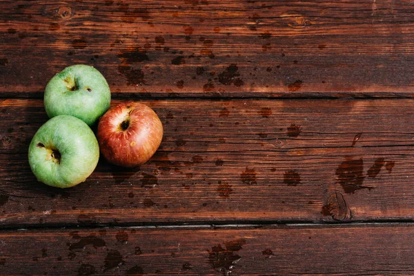 Three apples - two green and one red-lie on a wooden table. Fresh apples on a wood textured brown background, mahogany material. Fresh apples, autumn harvest.