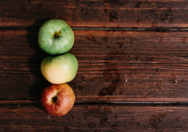 Three apples - green, yellow, and red-lie on a wooden table. Fresh apples on a wood textured brown background, mahogany material. Fresh apples, autumn harvest.