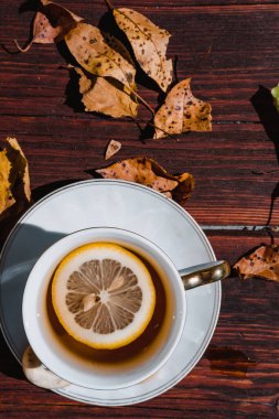 Tahta bir masanın üzerinde güzel beyaz bir çay fincanı ve yanında ağaçlardan düşen sarı sonbahar yaprakları var. Doğada limonlu ve şekerli siyah çay.