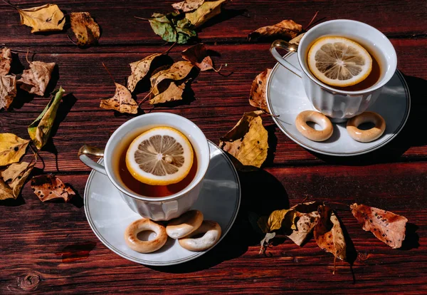 Beautiful white ceramic tea set on a wooden table, next to the fallen yellow autumn leaves from the trees. Black tea with lemon and sweets in nature, tea ceremony for two.