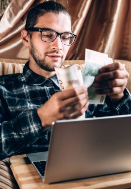 Mavi tişörtlü ve gözlüklü yakışıklı genç bir adam gümüş bir bilgisayarın başında çalışıyor ve elinde Avrupa kâğıdından para tutuyor. Rahat bir ev ortamında, çevrimiçi alışveriş yapan bir serbest çalışan için uzak çalışma.