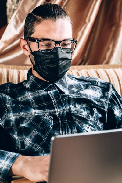 Diopter gözlüklü ve siyah koruyucu maskeli yakışıklı genç bir serbest çalışan evde gümüş bir dizüstü bilgisayarla çalışıyor. Karantina sırasında metnin basılması ve önemli işlerin yürütülmesi için uzak işlem.