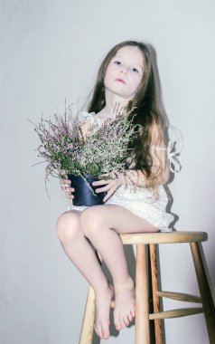 Beyaz bir arka plan üzerinde beyaz giysili bir tahta sandalyede oturan ve mor çiçekler bir pot holding beyaz kız