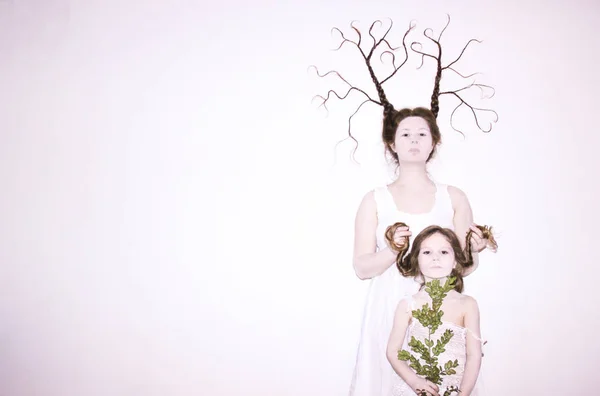 Mom and daughter in white dresses on a white background depict winter and spring, holding flowers and a twig with leaves. Hair braided in braids. Mom braid is in the form of wood