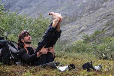 Genç bir erkek turist dinlenmek için crouches ve bacaklarını doğa zemin karşı yoga yaparken uzanıyor.