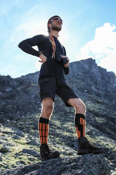 Young white male tourist in sportswear and boots standing on a rock in the mountains with a camera