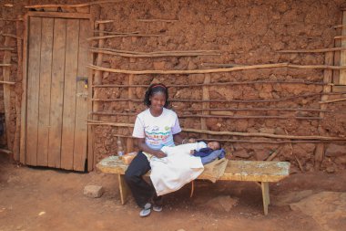 Kibera, Nairobi, Kenya - 13 Şubat 2015: Kucağında bir bebek tutan ve bir kil kulübe yakın bir bankta oturan beyaz bir T-shirt Afrika kadında