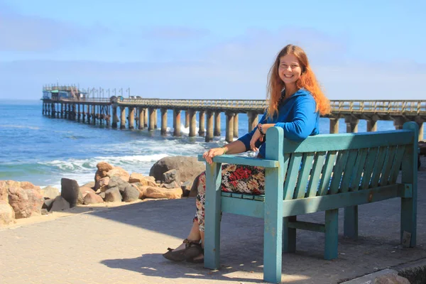 红头发快乐的白人白种人女孩坐在斯瓦科普蒙德 纳米比亚 大西洋沿岸的长凳上 在码头的背景上大笑 — 图库照片