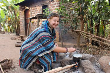 Mavi pançolu genç hippi kadın bir Afrika köyünde kamp ateşinde yemek pişiriyor.