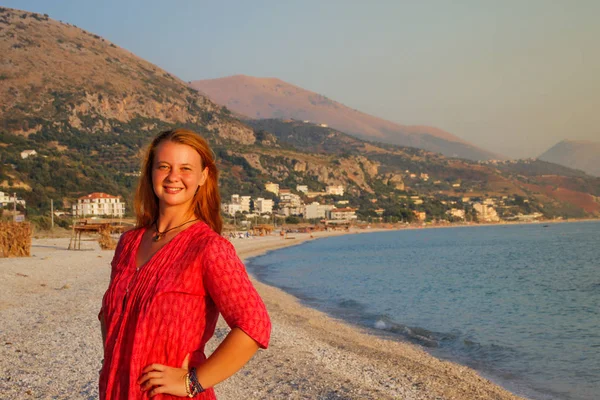 在地中海和阿尔巴尼亚海岸群山的背景下 一位身穿粉色长衫 红头发的美丽的白人年轻女子微笑着欣赏日落 — 图库照片