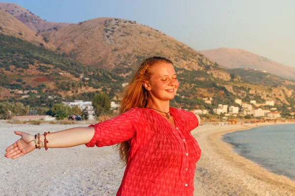 A beautiful young Caucasian woman in a pink blouse with red hair smiles and enjoys the sunset against the background of the Mediterranean Sea and the mountains on the coast of Albania.
