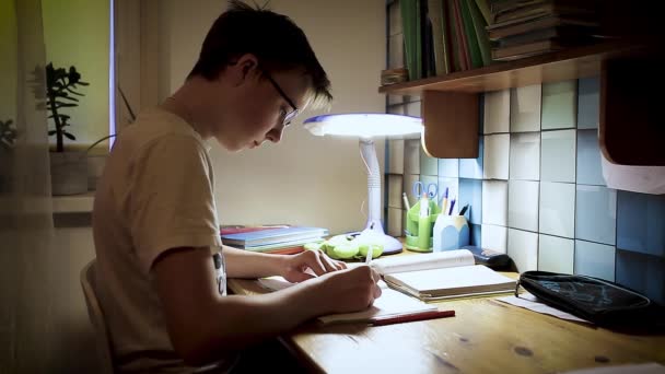 学校的男孩少年在眼镜学习做作业 男孩坐在桌旁 晚上在笔记上写字 — 图库视频影像