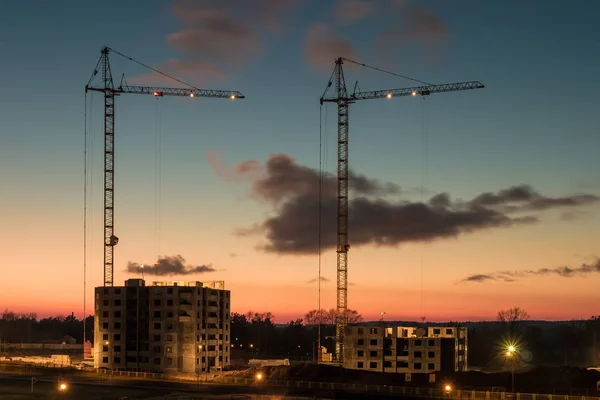 Silhouette tower cranes and unfinished multi-storey high buildings under construction at the sunset in desert on illuminated building site