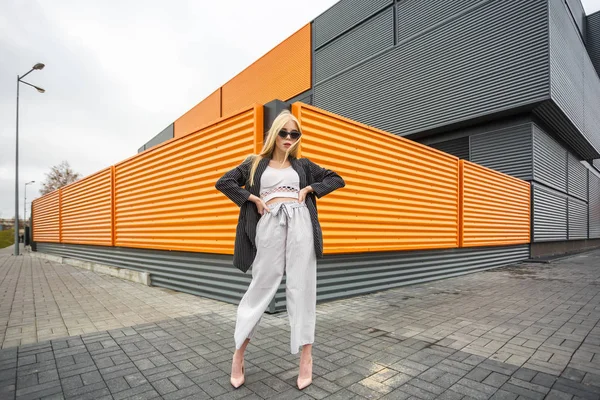 Gros plan portrait de mignon sourire petite fille modèle en veste rayée et lunettes de soleil posant près de gris ondulé rayé orange mur gris — Photo