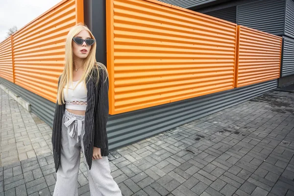 Gros plan portrait de mignon sourire petite fille modèle en veste rayée et lunettes de soleil posant près de gris ondulé rayé orange mur gris — Photo