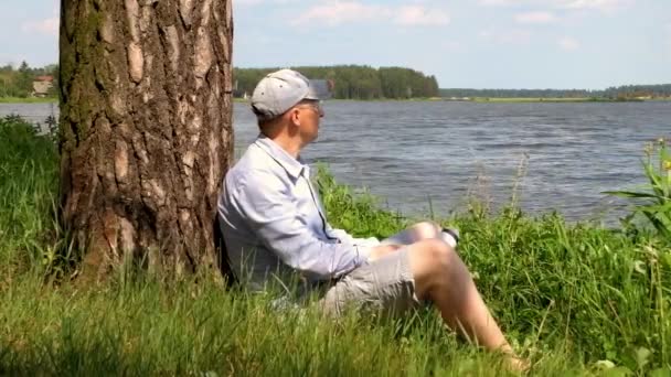 Muž Čepici Sedí Pod Stromem Břehu Jezera Pije Vodu — Stock video