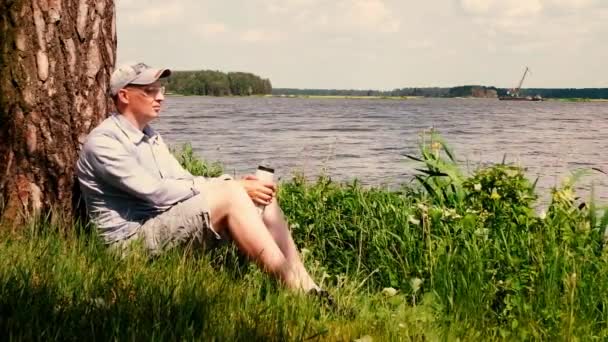 Man Cap Sitter Ett Träd Stranden Sjön Och Dricker Vatten — Stockvideo