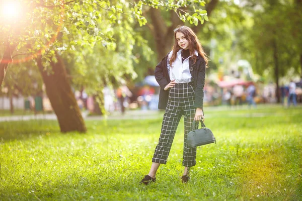 Portrait of little beautiful stylish kid girl with sunglasses and short plaid pants in city park on green forest background — Stock Photo, Image