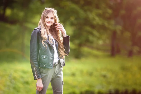 緑の森の背景に都市公園で革の黒いジャケットで長い髪を持つ小さな美しいスタイリッシュな背の高い子供の女の子の肖像画 — ストック写真