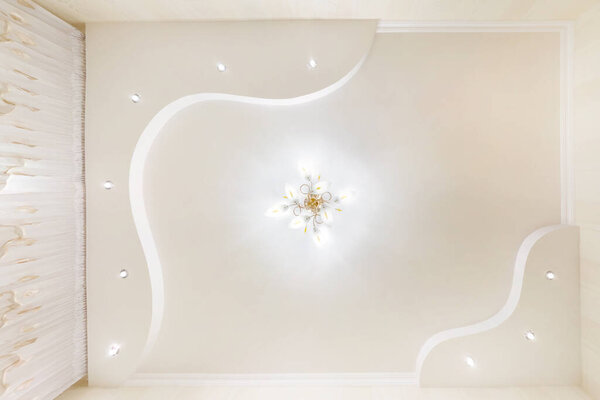 Растяжка потолка белой и сложной формы с галогенными пятнами ламп и гипсокартона строительства в пустой комнате в квартире или доме. Подвесный потолок . 
